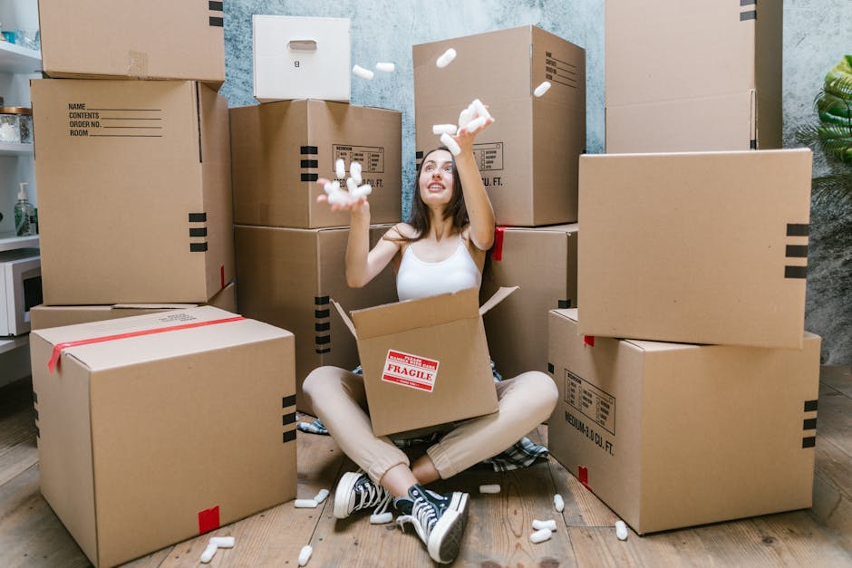 A young woman sitting on a wooden floor surrounded by numerous large cardboard boxes, some sealed with red tape, with packing materials such as Styrofoam packing peanuts falling from her raised hands.
